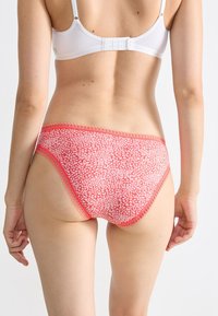 Vrouw in een witte beha en koraal-roze gestreepte slip met kant, gezien van achteren tegen een effen lichte achtergrond.