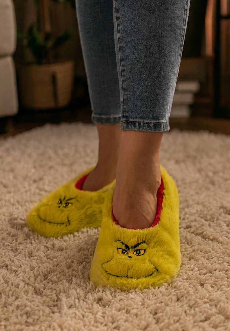 Pantofole di peluche gialle con rivestimento rosso, caratterizzate da un motivo con il volto del Grinch sulla parte anteriore. Texture morbida, forma arrotondata e aperte sul retro.