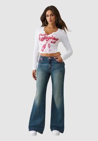 Biała koszula z długim rękawem z czerwonym nadrukiem graficznym, połączona z dżinsami z rozszerzanymi nogawkami, z efektem spranego denimu i kontrastującymi szwami. Białe trampki.