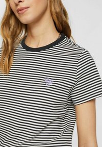 Gestreiftes T-Shirt in Schwarz und Weiß, mit rundem Ausschnitt und kurzen Ärmeln. Kleine lila Blumenstickerei in der Nähe der Brust.