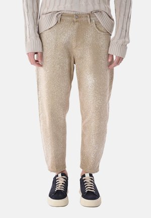 Pantaloni beige glitterati abbinati a sneakers nere e bianche con lacci, indossati con un maglione beige a maglia.