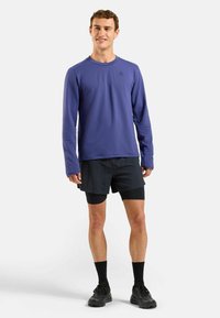 Lila Langarmshirt mit strukturiertem Muster, kombiniert mit schwarzen Shorts. Das Outfit hat ein Logo auf der Brust und schwarze Schuhe.