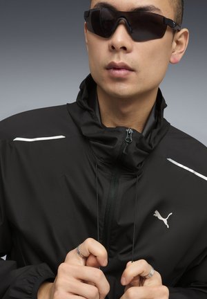 Veste noire à fermeture éclair avec un col montant, ornée d'accents blancs et d'un logo argenté. Portée avec des lunettes de soleil foncées, affichant un design minimaliste.