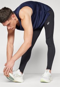 Mand strækker sig, iført en marineblå ærmeløs shirt, sorte leggings og hvide sportssko med grønne accenter. Tekstureret stof med synlige sømme.