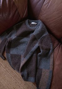 Grau und braun gemusterter Pullover aus weich strukturiertem Wollmaterial, mit Rundhalsausschnitt und überschnittenen Schultern, auf einem braunen Ledersofa ausgelegt.
