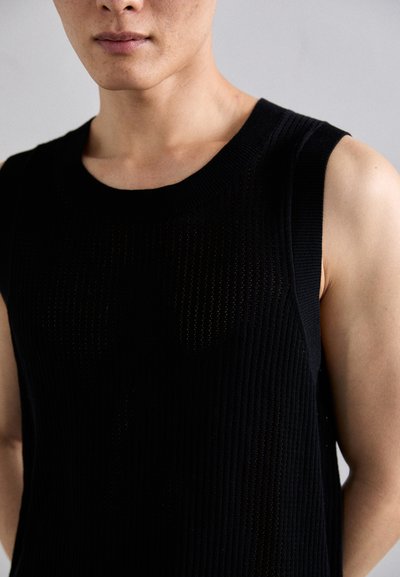 Man wearing sleeveless black ribbed knit top with subtle transparent pattern against light gray background.