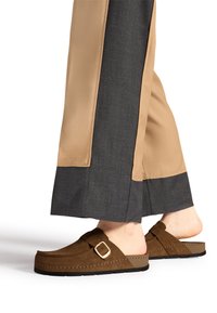 Persona che indossa scarpe slip-on in suede marrone con dettaglio in oro e pantaloni a gamba larga bicolore beige e grigio scuro.