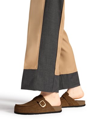Person iført brune ruskind slip-on sko med guld spænde og to-farvede beige og mørkegrå brede bukser.