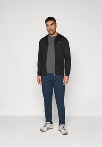 Black zip-up hoodie with yellow logo, gray t-shirt, and navy joggers; paired with white sneakers featuring gray accents and a textured sole.