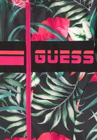 Tessuto nero con foglie tropicali verdi e bianche e fiori rosa, cerniera verticale fucsia brillante e logo 'GUESS' in fucsia su banda nera.