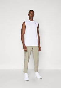 White sleeveless athletic shirt with black logo, paired with light beige tapered pants and white sneakers, standing on a plain background.