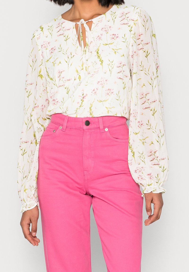 Femme portant un chemisier blanc à fleurs à manches longues avec un nœud à l’avant et un pantalon taille haute rose vif sur un fond uni.