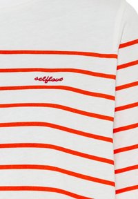 Camisa de manga larga blanca con amplias rayas horizontales naranjas. Presenta bordado "selflove" en rojo en la zona del pecho. Tejido de algodón.