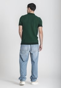 Camisa polo texturizada verde con acentos negros, combinada con jeans holgados azul claro y zapatos color crema, vista desde atrás.