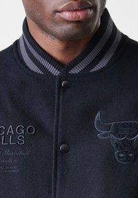 Close-up de uma pessoa a usar uma jaqueta preta dos Chicago Bulls com gola riscada, botões pretos e o nome da equipa e o logótipo do touro bordados.