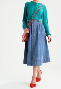 Blusa de manga larga en color azul verdoso combinada con una falda de mezclilla azul con botones. Un bolso rojo y unos zapatos rojos de tipo slip-on completan el conjunto.