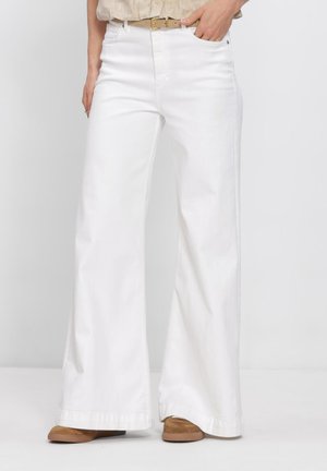 Frau trägt hoch taillierte, weit geschnittene weiße Hose mit beigem Gürtel und braunen Schuhen, steht vor einem einfarbigen Hintergrund.