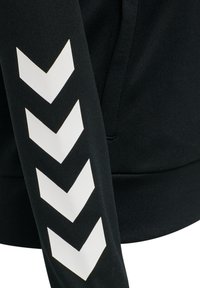 Czarny rękaw od bluzy z trzema dużymi białymi chevronami. Tkanina ma gładką fakturę, z ribbowanym mankietem i bezszwowym wzornictwem.