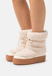 Buty z kremowego, pluszowego materiału o puszystej fakturze, z okrągłym noskiem, grubą brązową podeszwą i związanymi sznurówkami wokół kostki.