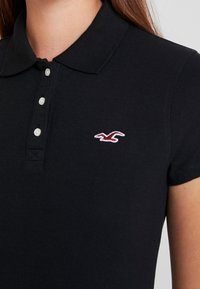 Svart polotröja i bomull, med krage, tre knappar fram och broderad logotyp i röd och vit på bröstet.