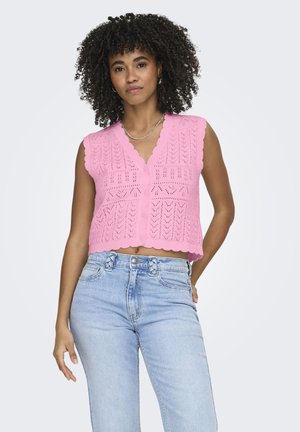 Donna con capelli ricci che indossa un crop top lavorato a maglia rosa senza maniche e jeans azzurri a vita alta, in piedi con una mano dietro la schiena.