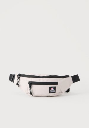 Lichtbeige heuptas met zwarte ritsen, verstelbare band en Champion-logopatch op het voorvak.