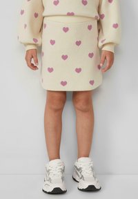 Bambina che indossa una gonna a maglia color crema e un maglione con motivi a cuore rosa, sneaker bianche e grigie, in piedi su uno sfondo semplice.