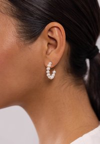 Ohrringe mit Hoops aus weißen Perlen und goldenen Akzenten, die ein gebogenes Design aufweisen, das durch das Ohr läuft. Strukturierte Oberfläche mit Glanz.