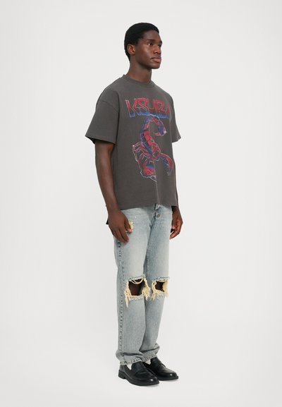 Graues Grafik-T-Shirt mit Skorpion-Design, kombiniert mit hellblauen, verwaschenen Jeans mit fransigen Säumen und schwarzen Schlupfschuhen.