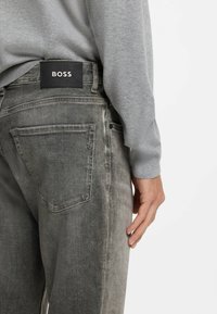 Szare dżinsy z denimu o teksturowanym wykończeniu, tradycyjne kieszenie z tyłu oraz czarna naszywka z logo na pasku. Widoczny casualowy, szary sweter.