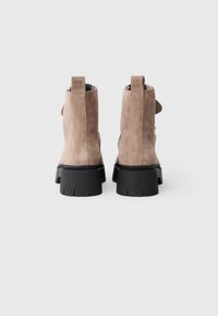Botoșei din suede la gleznă, de culoare maro deschis, cu o talpă groasă din cauciuc negru. Prezintă un logo embosat pe toc și o buclă pentru o îmbrăcare ușoară.