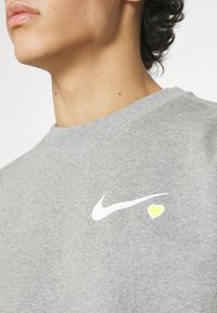Heathergrå sweatshirt, blandning av bomull, rund hals, med en vit Nike-logga och en gul hjärtan detalj på bröstet. Slät textur.