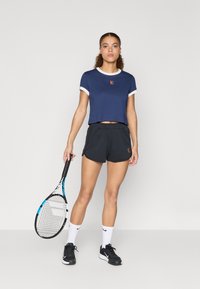 Ναυτικό κροπ τοπ με λευκή επένδυση, λεπτομέρεια λογότυπου, μαύρη βερμούδα και μαύρα αθλητικά παπούτσια. Κρατάει ένα ρακέτα τένις, φορώντας λευκές κάλτσες. Μινιμαλιστικός σχεδιασμός.