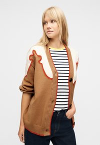 Ξανθιά γυναίκα που φοράει καφέ και κρεμ πουλόβερ με κόκκινο περιθώριο πάνω από ένα μαύρο-λευκό ριγέ πουκάμισο και σκούρο τζιν, κοιτάζοντας προς τα αριστερά της.