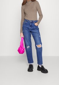 Aangesloten bruine coltrui, hoogtijdse blauwe jeans met gescheurde details, en een felroze satijnen tas; zwarte plateau schoenen; neutrale achtergrond.