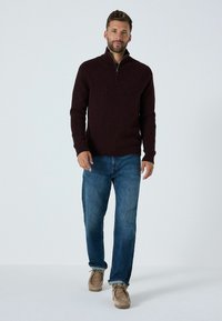 Weinroter Pullover mit Reißverschluss aus strukturiertem Strickstoff, kombiniert mit blauen Jeans und beigen Schuhen. Moderner Schnitt mit langen Ärmeln.
