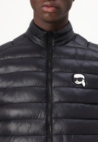Melna pūkaina veste ar horizontālu šuvumu dizainu, augstu apkakli un priekšējo rāvējslēdzēju. Iezīmējas ar izteiksmīgu logo uz kreisā krūts.