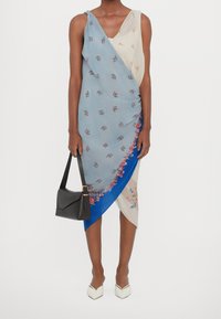 Robe asymétrique bleu et crème, ornée de motifs floraux, avec une coupe drapée et un accent bleu contrastant. Sac à main noir et chaussures blanches.