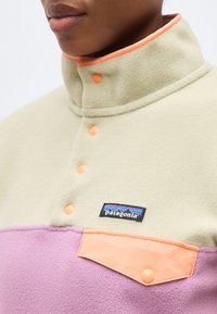 Κοντινό πλάνο ενός ατόμου που φοράει μπεζ και μοβ fleece Patagonia με πορτοκαλί κουμπιά και καπάκι στην τσέπη στο στήθος.