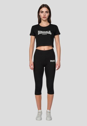 Kvinde står iført sort Lonsdale cropped t-shirt og matchende capri-leggings med hvide sneakers på grå baggrund.