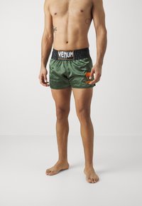 Calções de boxe verde em satin com faixa elástica na cintura. Apresenta o logótipo "VENUM" em preto na faixa e o logótipo laranja no lado esquerdo.