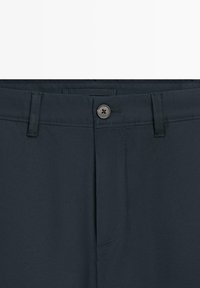 Primo piano della vita di pantaloni blu scuro con chiusura a bottone, passanti per cintura e dettaglio della cucitura frontale.