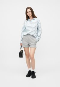 Νεαρή γυναίκα με ανοιχτό μπλε πουκαμίσα με ρίγες και μακριά μανίκια, γκρι σορτς, λευκές κάλτσες, μαύρα αθλητικά παπούτσια, κρατώντας μια μαύρη τσάντα, στέκεται πάνω σε λευκό φόντο.