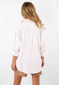 Chemise blanche légère à manches retroussées, en tissu texturé, avec un détail d'ouverture au dos. présente une petite étiquette de marque près de l'épaule.