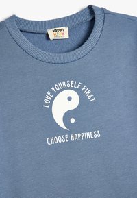 Sudadera de algodón azul con cuello redondo con acanalado. Presenta un diseño de yin-yang en blanco y el texto "Ámate a ti mismo primero, elige la felicidad."