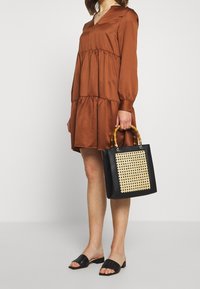 Brun klänning i flera lager med långa ärmar, svart väska med vävd framsida och bambuhandtag, samt platta svarta sandaler med en texturerad design.