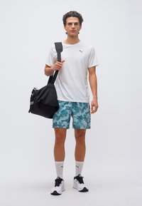 Νέος άνδρας που φοράει λευκό t-shirt Puma, μπλε καμουφλαρισμένο σορτς, λευκές κάλτσες Puma και λευκά αθλητικά παπούτσια, κρατώντας μια μαύρη τσάντα γυμναστηρίου στον ώμο του.