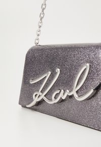 Geantă dintr-un gri metalic cu sclipici, cu o curea de lanț argintie; prezintă inscripția embossată "Karl" din metal lucios pe față. Design compact, rectangular.