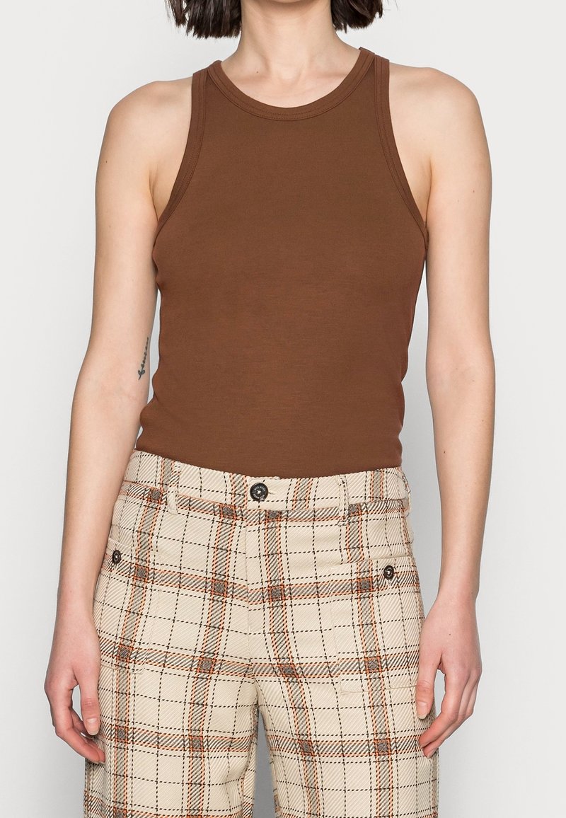 Femme portant un haut marron sans manches et un pantalon taille haute à carreaux crème avec des lignes orange et noires.