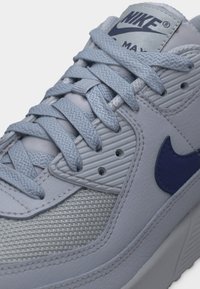 Nærbilde av en lysegrå Nike Air Max-sneaker med teksturert mesh, sydd skinn, lisser og mørkeblå Nike Swoosh-logo.